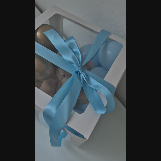 Kleine hellblaue und goldene Luftballons in einer weißen Geschenkbox. Mit Teddybär in der Box und hellblauem Seidenband mit Schleife um die Box herum. 