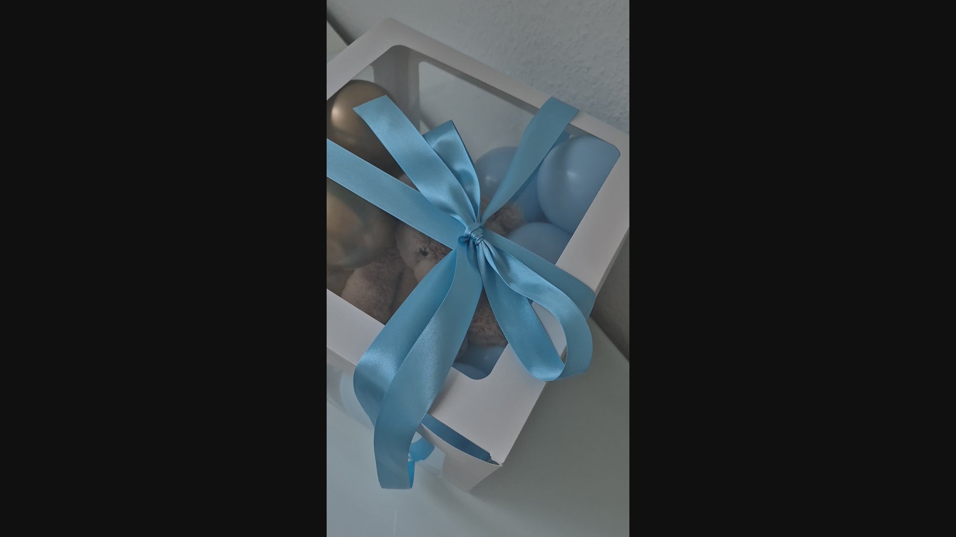 Kleine hellblaue und goldene Luftballons in einer weißen Geschenkbox. Mit Teddybär in der Box und hellblauem Seidenband mit Schleife um die Box herum. 